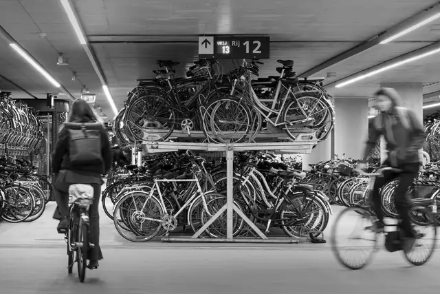 Cyclistes traversant un grand parking à vélos couvert, aux râteliers à deux étages remplis de vélos